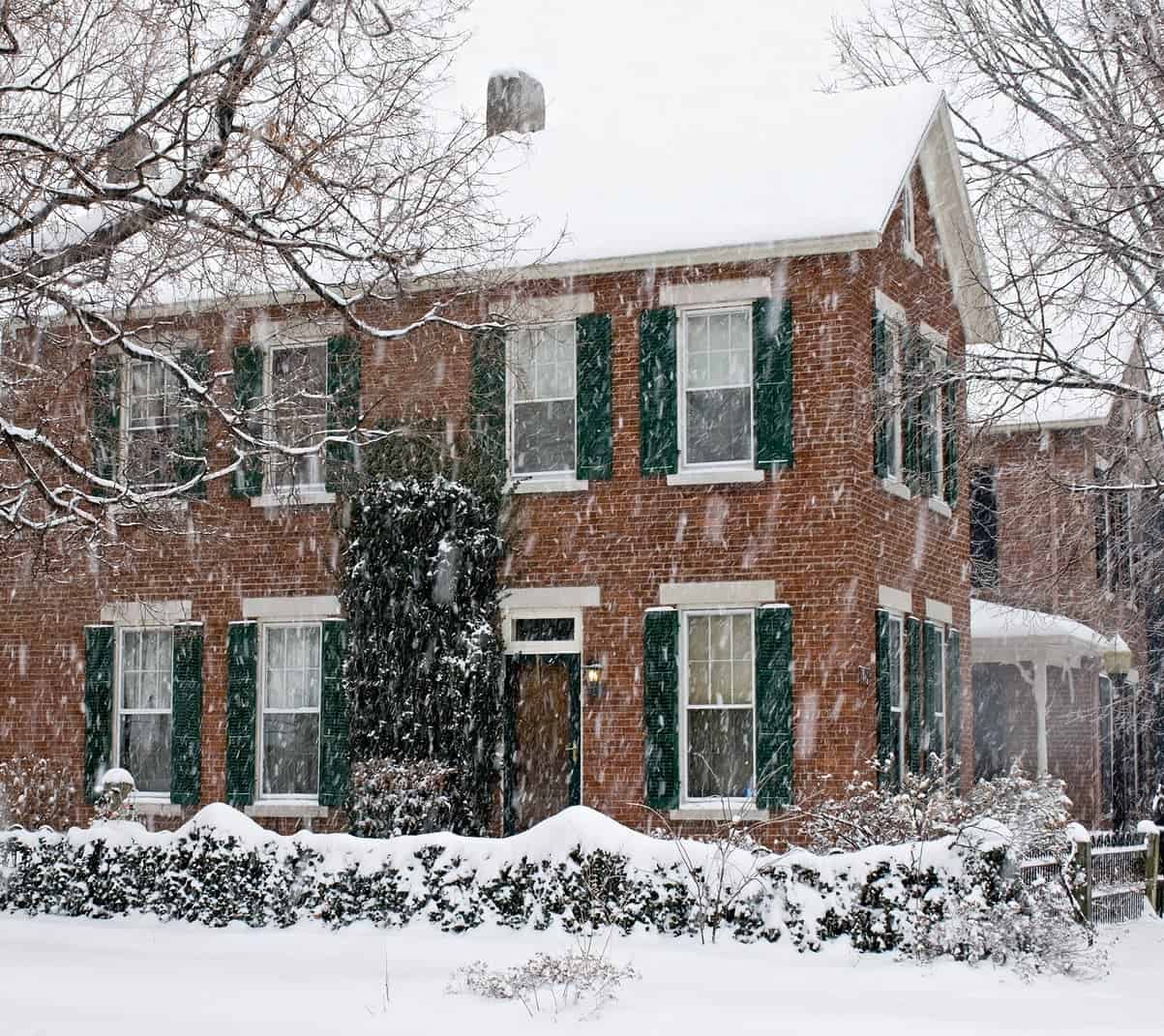 Historic brick house during snowfall with green shutters and snow-covered yard.