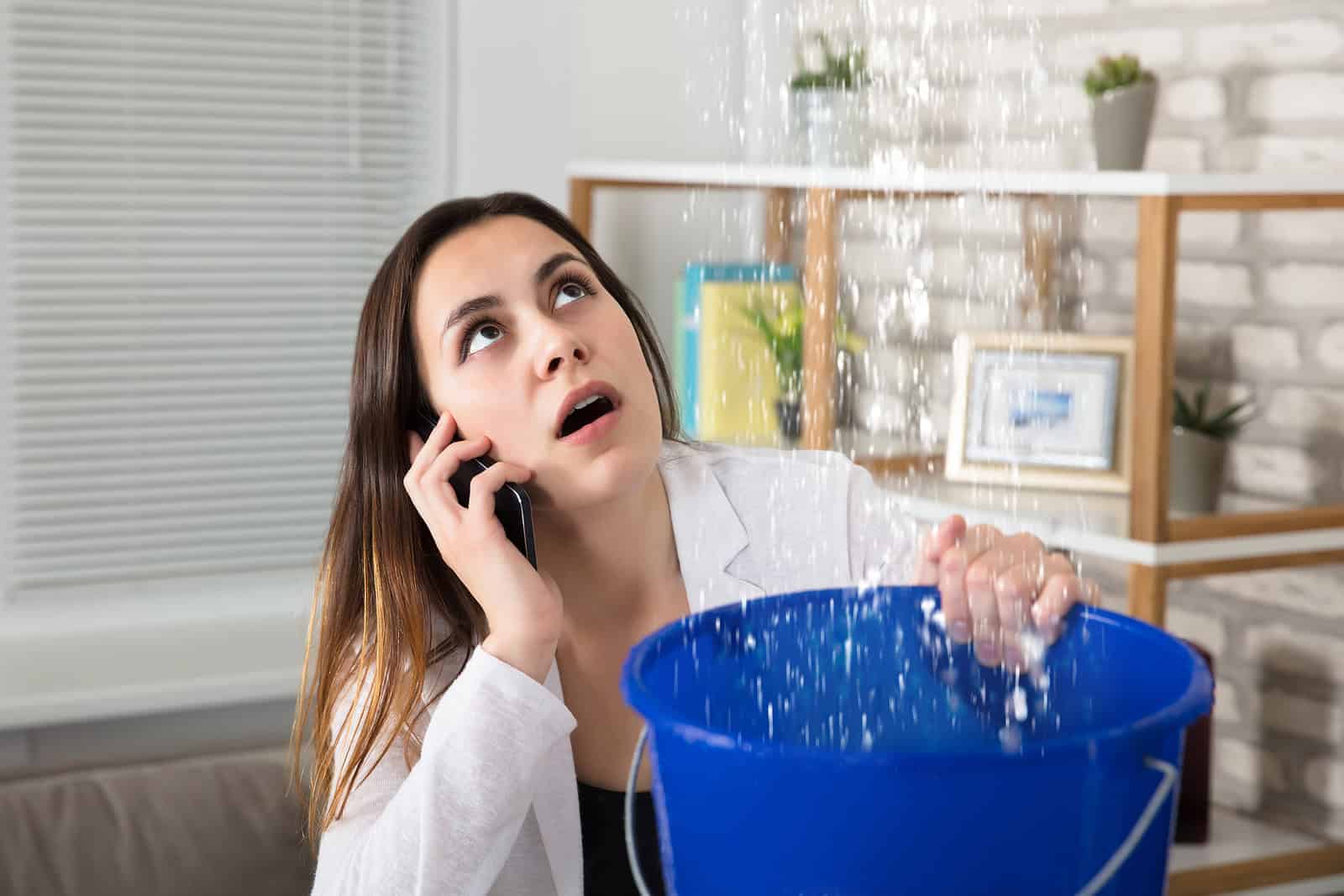 Woman calling for help while cleaning a flooded room with a bucket.