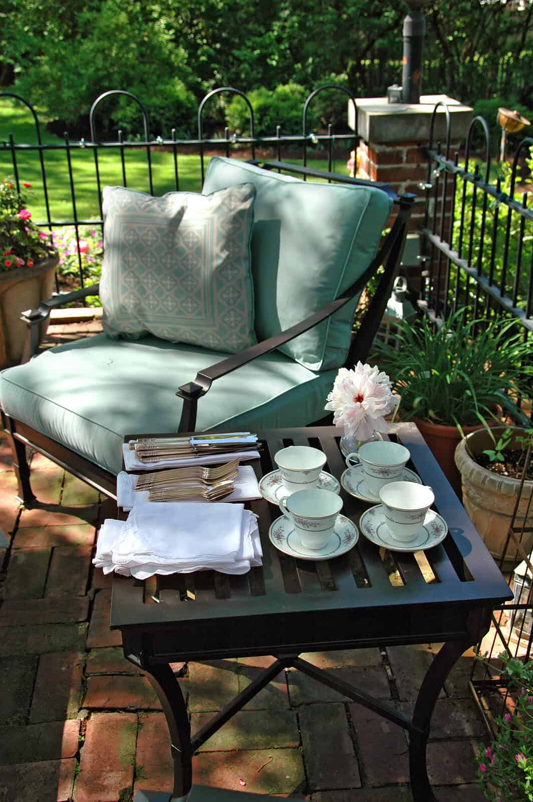 A cozy outdoor patio with a vintage-style cushioned chair, tea set, and potted plants in a lush gard.