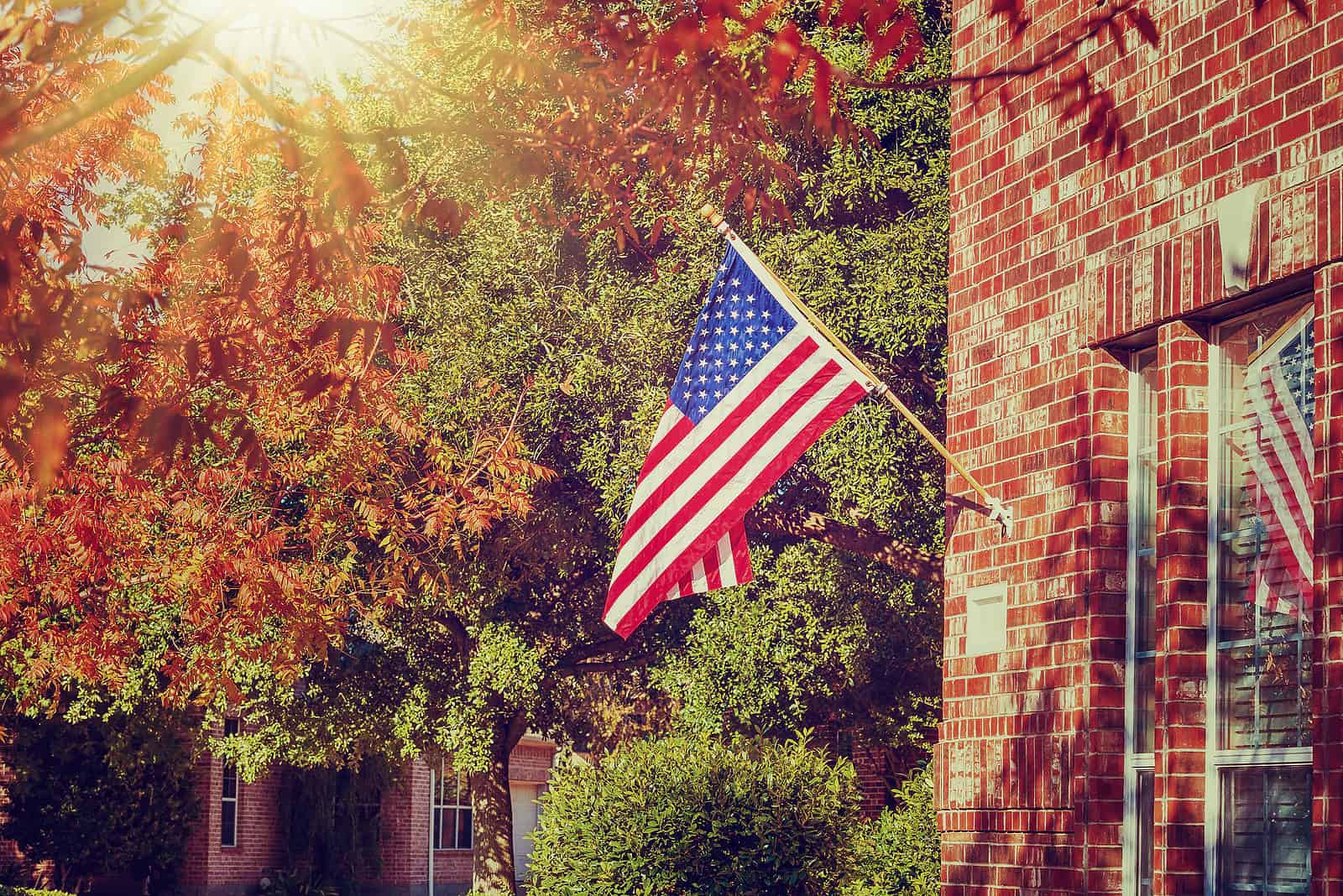 American flag hanging on brick building with autumn trees in background.