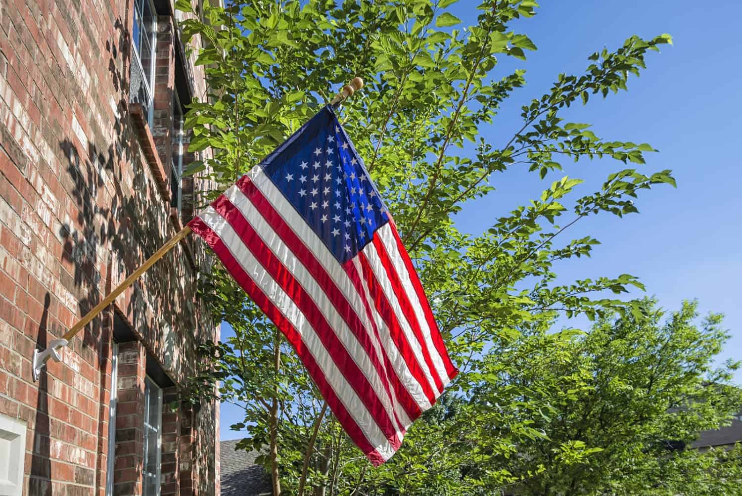 American flag hanging on a historic brick building with green trees and blue sky in the background.