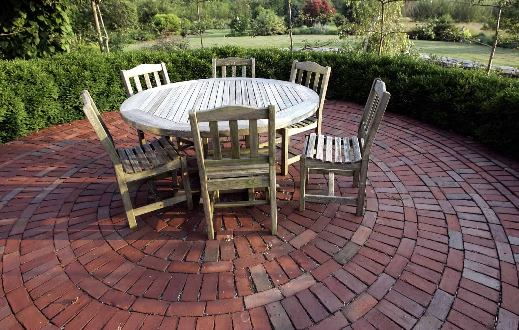 Rustic outdoor patio with a round wooden table and six chairs on a brick circular surface.