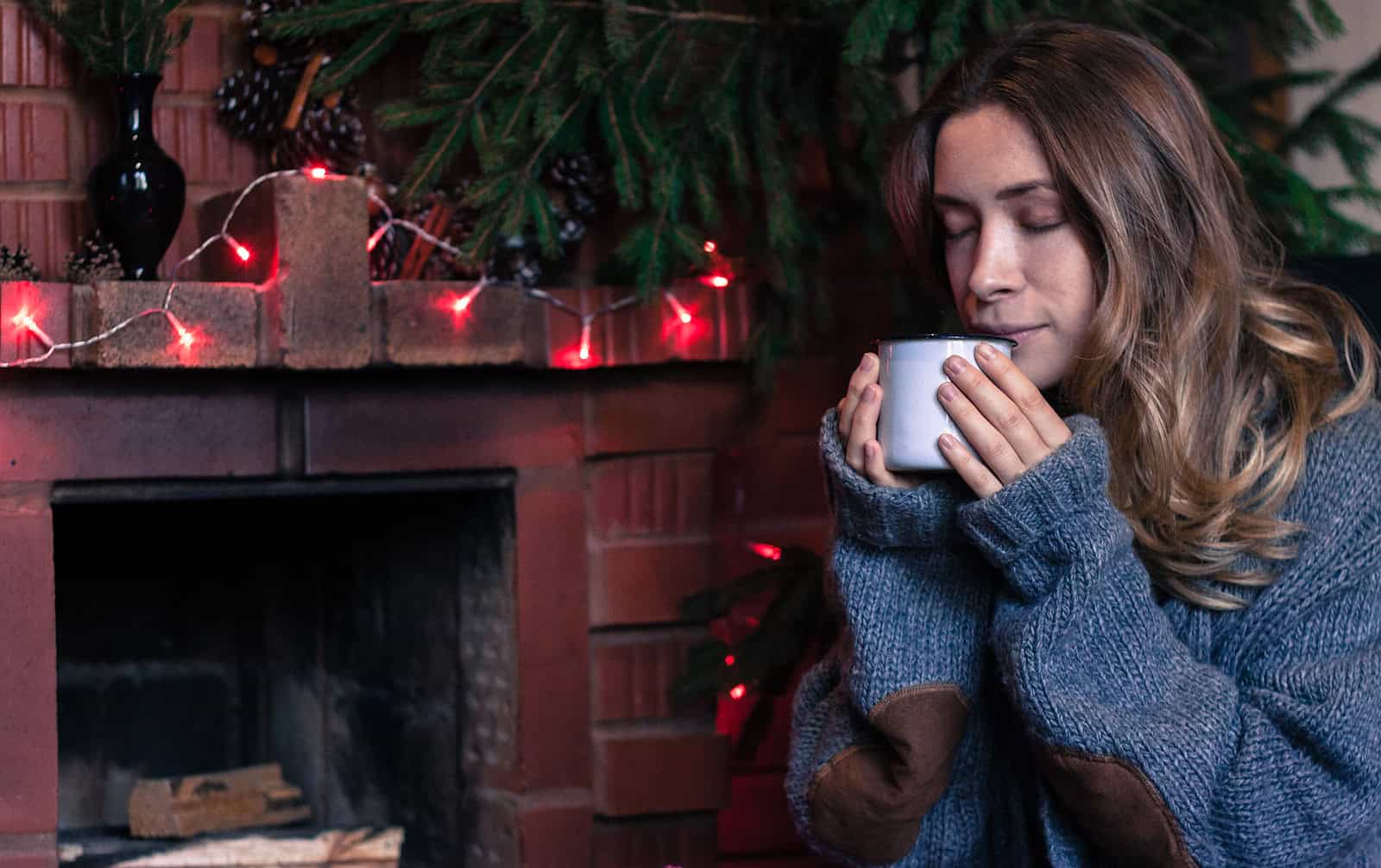 A woman enjoying a warm drink near a decorated fireplace with Christmas lights and greenery.