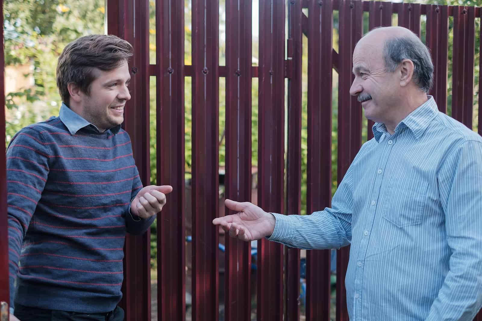 Two men talking outdoors near a wooden fence, discussing masonry work.
