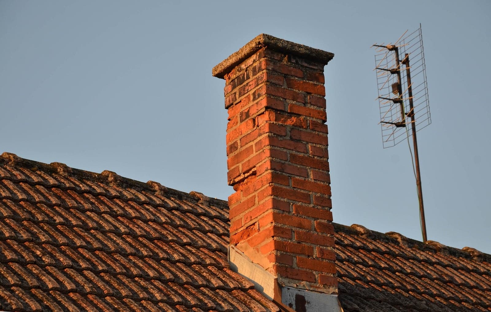 Close-up of a brick chimney on a tiled roof with an antenna in the background.