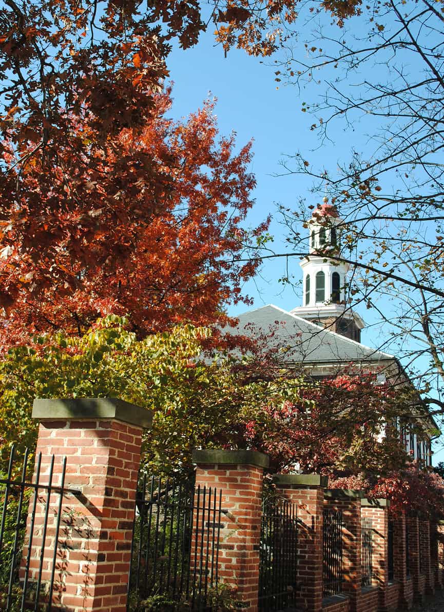 Historic brick church with a white steeple, surrounded by autumn trees and a wrought iron fence.